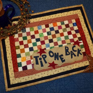 May include: A quilted table runner with a patchwork design in red, white, and blue. The runner has a border of red and white gingham and a navy blue trim. The text "Tip One Back" is embroidered on the runner in blue.