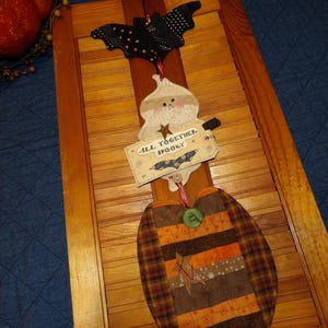 May include: A Halloween-themed wooden decoration featuring a bat, a ghost, and a pumpkin. The bat is black with white polka dots. The ghost holds a sign that reads "ALL TOGETHER, SPOOKY". The pumpkin is made of fabric with orange, brown, and tan stripes.