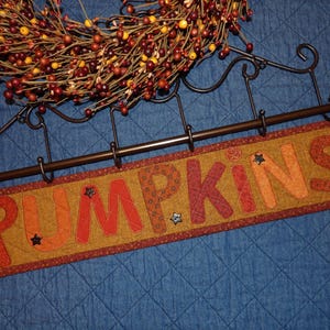 May include: A rectangular, quilted sign with the word "PUMPKINS" in orange and red letters. The sign is on a dark metal rod with hooks, and a fall wreath is above it. The sign has star accents.