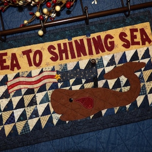 May include: A rectangular quilted wall hanging featuring a brown whale appliqué on a blue and cream triangular patterned background. The text SEA TO SHINING SEA is appliquéd in dark red letters across a cream banner at the top with a small flag.