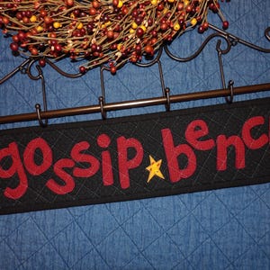 May include: A black quilted sign with the words "gossip bench" in red letters and a yellow star. The sign hangs from a decorative metal rod with hooks, and is topped with a fall-themed wreath.