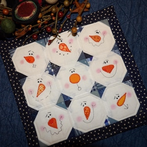 May include: A white quilted fabric square with a blue border and white stars. The square is divided into nine sections, each featuring a different snowman face with orange noses and black eyes.