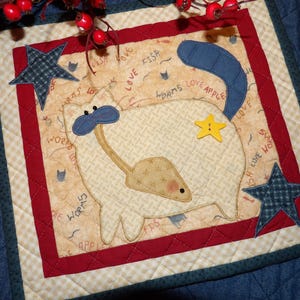 May include: A quilted square featuring a whimsical cat design. The cat is cream-colored with blue and yellow accents, holding a mouse. The background is beige with red and blue borders, and star-shaped embellishments. Text includes "worms love fish".
