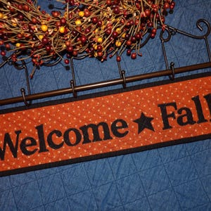 May include: A rectangular orange quilted fabric sign with black trim and the black text "Welcome Fall" featuring a star between the words. The sign hangs from a dark metal rack with five hooks, positioned below a wreath of red and yellow berries.