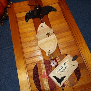 May include: Halloween-themed hanging decoration featuring a black glitter bat, a ghost-shaped applique, and a pumpkin-shaped applique. The pumpkin has a tag that says "You've been boo'd". The decoration is made of fabric and has button and star accents.