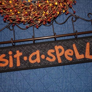 May include: A black sign with the words "sit-a-spell" in orange letters. The sign is hanging from a decorative metal rod with hooks, and a fall wreath is above it.