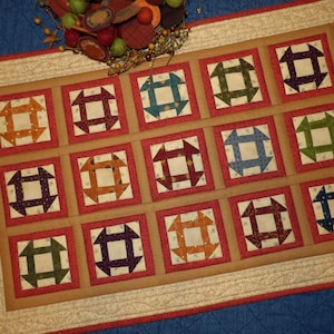 May include: A beige and red quilted table runner with a repeating pattern of squares with a geometric design in various colors, including red, green, blue, and purple.