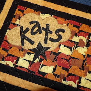 May include: A black and yellow quilted doormat with a brown cloud-shaped applique that says "Kats" in black lettering. The background of the doormat is a pattern of orange, brown, and white cats.