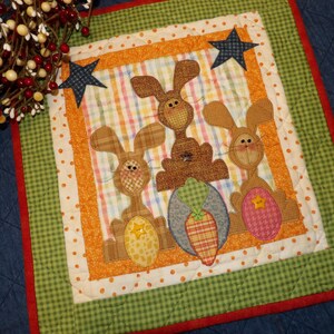 May include: A quilted Easter-themed wall hanging featuring three brown bunnies with patterned ears and bodies. The bunnies are centered on a plaid background, surrounded by orange and green borders, and accented with star shapes.