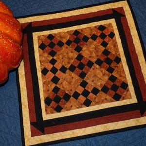 May include: A square quilted placemat with a central pattern of black and brown squares. The border features black, red, and cream-colored stripes. A large orange pumpkin is partially visible on the left.