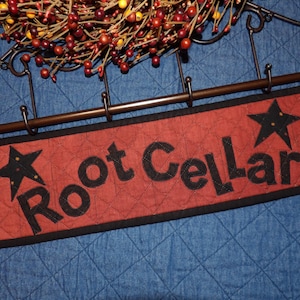 May include: A rectangular quilted fabric sign with a rust-colored background and black border. The text Root Cellar is appliqued in black letters, flanked by two black stars. The sign hangs from a dark metal rack with hooks and decorative scrollwork.