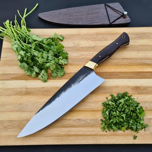 May include: A handcrafted chef's knife with a dark wood handle, brass bolster, and a polished blade. The knife rests on a bamboo cutting board next to fresh cilantro and a wooden sheath. A culinary tool for chopping and slicing.