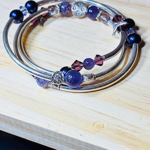 May include: Three silver bangles adorned with a variety of beads. The beads include dark blue, purple, and clear crystal beads, along with silver decorative beads. The bracelets are stacked on top of each other.