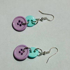 Puede incluir: Un par de pendientes hechos a mano con un botón grande de color lavanda, un botón pequeño turquesa y un gancho plateado. Los pendientes están conectados por pequeños anillos plateados. Los botones tienen cuatro agujeros.