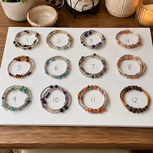 May include: A collection of twelve multi-strand wire wrap bracelets displayed on a white board, each numbered one through twelve. The bracelets feature various colorful beads, including amber, turquoise, purple, orange, and brown tones on metal wire.
