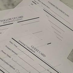 May include: A stack of white paper forms with black text and lines. The forms include "Follow Up Card," "Communication Log," and "Guest Card" with fields for information. The forms are on a light-colored surface.