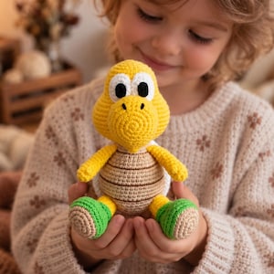 May include: A yellow crocheted turtle-like creature with a tan shell, white eyes with black pupils, and green feet. The figure features a textured yarn construction with a rounded head, small arms, and a distinctively patterned tan belly section.