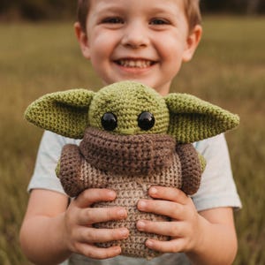 May include: A crocheted plush toy resembling a character with large ears and black eyes. The toy is mainly green, brown, and beige, and is held by a person. The background is blurred, suggesting an outdoor setting.