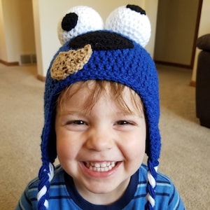 May include: A blue crocheted hat with white and black accents, resembling a cartoon character. The hat has two white, round shapes with black spots on top, and a brown cookie-shaped patch. The hat has blue and white braided ties.