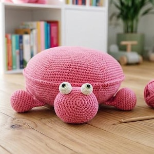 Op de afbeelding: Een gehaakt schildpadkussen in roze kleur met een afgerond schild, een kleine kop met twee witte ogen met zwarte pupillen en vier korte pootjes. Het item is gemaakt van garen en ligt op een houten ondergrond naast een bol bijpassend roze garen.