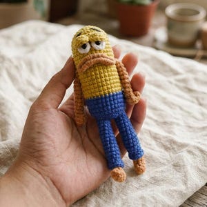 Könnte beinhalten: Eine kleine, handgefertigte Häkelpuppe mit einem gelben, zylindrischen Kopf, einem mürrischen Gesichtsausdruck, hängenden Augen und einem beigefarbenen Mund. Die Puppe hat einen gelben Oberkörper, eine blaue Hose sowie beigefarbene Arme und Füße.