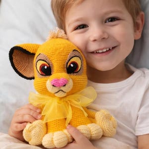 May include: A crocheted Simba plush toy in shades of yellow, tan, and pink, with a yellow ribbon. The toy has large, expressive eyes and is held by a smiling child. The plush toy is a handmade item.