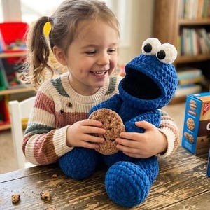 May include: A blue crocheted Cookie Monster plush toy holding a brown crocheted cookie. The plush has large white eyes and a wide open mouth. The toy is being held by a smiling child wearing a striped sweater.