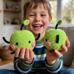May include: Two round, lime green crocheted plush toys with black stems and small green leaves. Each toy features black embroidered eyes, pink crocheted cheeks, and small black limbs. The toys have a textured, yarn-based surface.