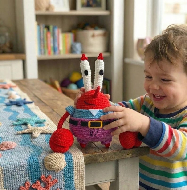May include: A crocheted Mr. Krabs plush toy. The toy has a red head, white eyes, and a blue shirt with a purple body. The plush is held by a child. A blue and white table runner with sea-themed decorations is in the foreground.