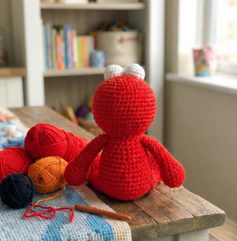 May include: A handmade red crochet doll with white eyes and limbs. Several balls of yarn in red, orange, and black are next to the doll, along with a crochet hook. The doll is on a wooden table.
