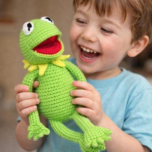 Op de afbeelding: Een handgemaakte groene gehaakte kikkerpop met witte ogen, een rode mond en een gele kraag. Het pluche speelgoed wordt vastgehouden door een persoon die een blauw shirt draagt. De kikker heeft lange armen en benen.