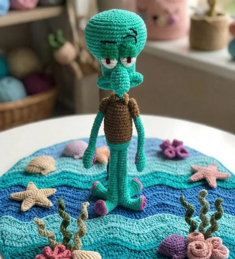 May include: A handmade crochet Squidward doll in teal, brown, and pink. The doll is posed on a blue and teal crocheted blanket with starfish, seashells, and coral accents. The doll has a grumpy expression.