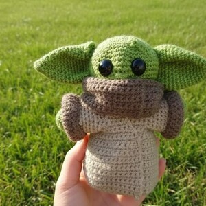 May include: A handmade crochet doll of a character with large ears, large black eyes, and a beige body. The doll has a green head and ears, a brown neck and arm coverings, held against a green grassy background.