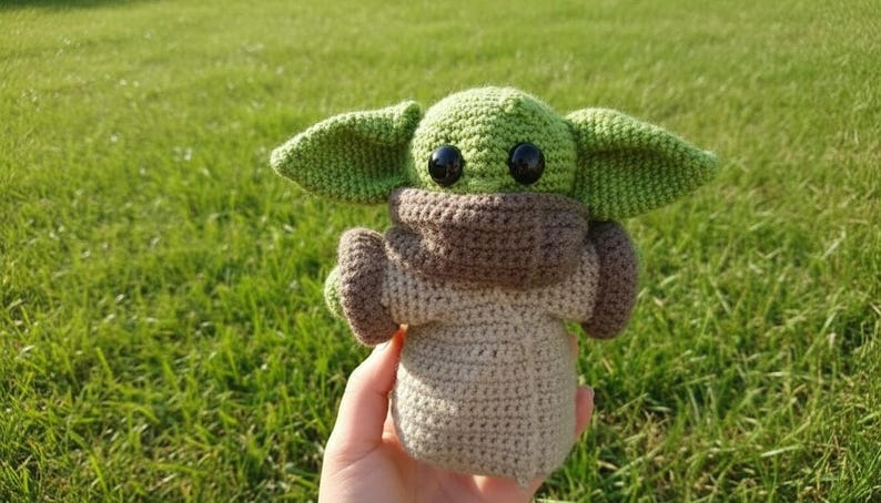 May include: A handmade crochet doll of a character with large ears, large black eyes, and a beige body. The doll has a green head and ears, a brown neck and arm coverings, held against a green grassy background.