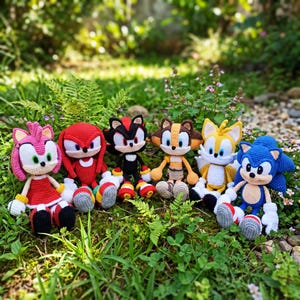 May include: Six handmade crochet plush toys representing characters from the Sonic the Hedgehog series, including Amy Rose, Knuckles, Shadow, Sticks, Tails, and Sonic, all arranged in a row on a patch of green grass and small flowers.