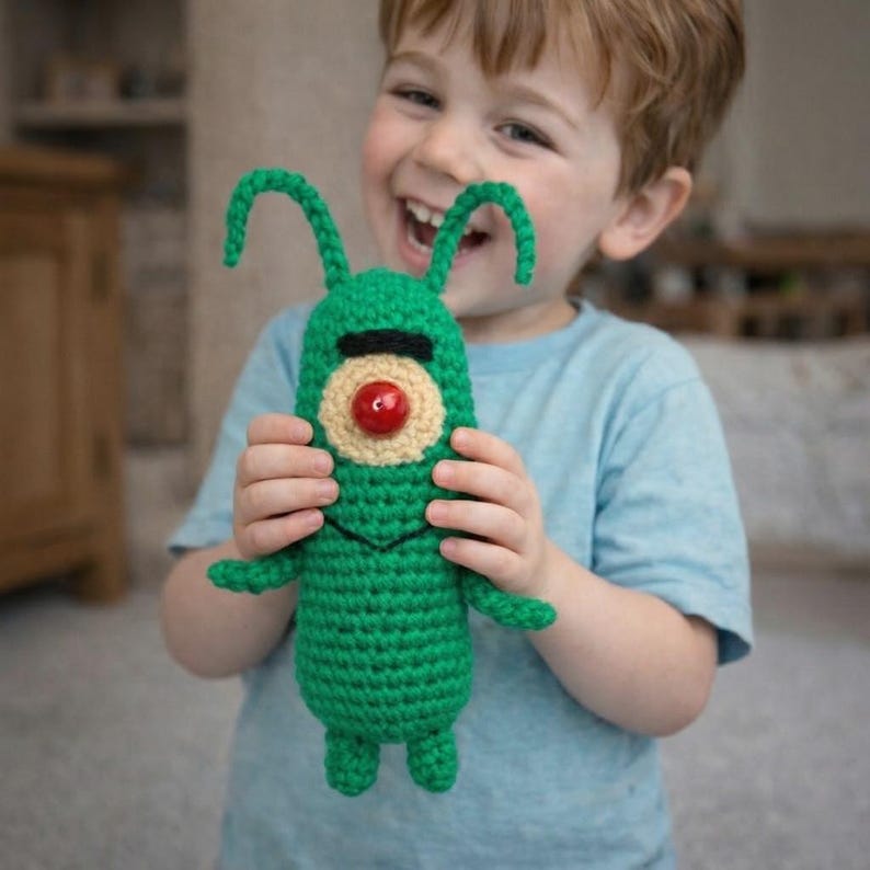 May include: A handmade green crochet Plankton toy with a black eye, beige eye outline, and a red nose. The toy has two green antennae and small arms and legs. The toy is held by a child.