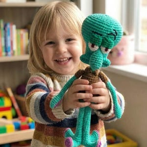 Könnte beinhalten: Gehäkelte Squidward-Puppe in Türkis mit braunen und lila Akzenten. Das lächelnde Kind trägt einen gestreiften Pullover. Die Puppe hat einen grimmigen Gesichtsausdruck und lange, schlanke Beine. Handgefertigt.
