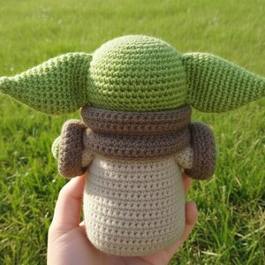 May include: A crocheted doll featuring a character with large green ears, a brown collar, and a beige body. The doll is held against a backdrop of green grass. The doll is a popular character from a science fiction franchise.