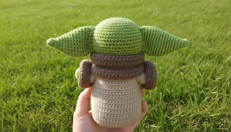 May include: A crocheted doll featuring a character with large green ears, a brown collar, and a beige body. The doll is held against a backdrop of green grass. The doll is a popular character from a science fiction franchise.