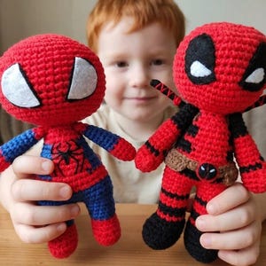 Könnte beinhalten: Zwei handgefertigte Häkelpuppen von Superhelden. Einer ist Spider-Man, mit einem rot-blauen Anzug, weißen Augen und einem schwarzen Spinnenemblem. Der andere ist Deadpool, in einem rot-schwarzen Anzug mit weißen Augen und einem braunen Gürtel.