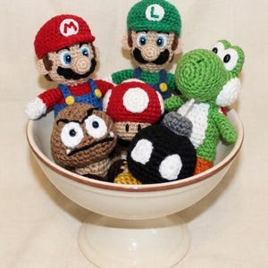 Könnte beinhalten: Eine Sammlung handgefertigter Häkelfiguren von Charakteren aus der Super Mario-Videospiel-Franchise. Die Schüssel enthält Mario, Luigi, Yoshi, einen Goomba, einen Pilz und eine Bob-omb. Die Figuren befinden sich in einer weißen Schüssel mit Sockel.