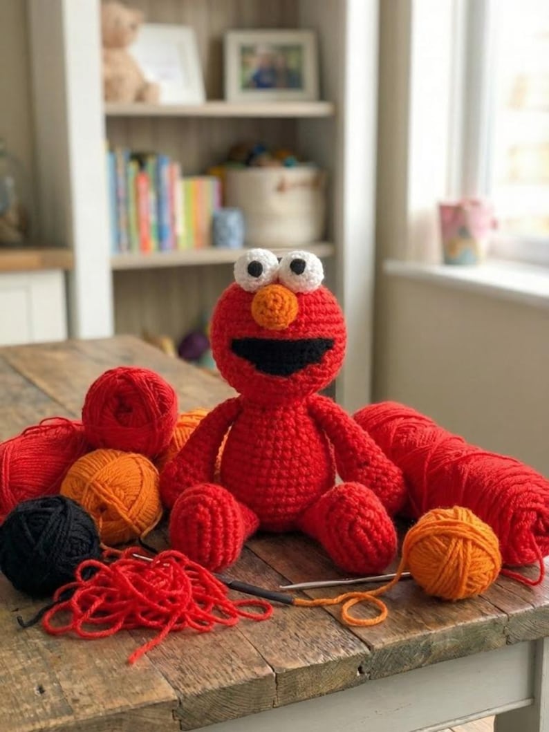 Op de afbeelding: Een handgemaakte rode gehaakte figuur met witte ogen, een oranje neus en een zwarte mond zit op een houten tafel. Verschillende bollen rood, oranje en zwart garen omringen het figuur, samen met haaknaalden. Een knutselproject.