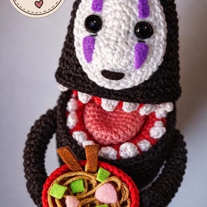 May include: A crocheted figure with a black body, a white face featuring purple markings, and a wide open mouth with white teeth and a pink tongue. The figure holds a red bowl containing brown yarn noodles, green felt squares, and pink felt pieces.