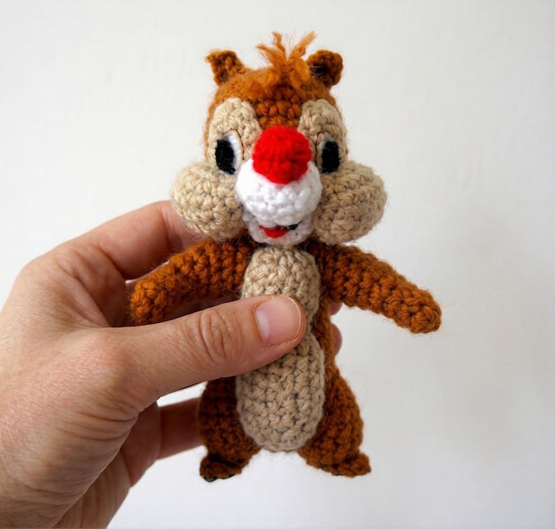 May include: A handmade crochet Chipmunk doll in brown, tan, white, and red. The doll features a red nose, white muzzle, and black eyes. The Chipmunk is held in a hand.