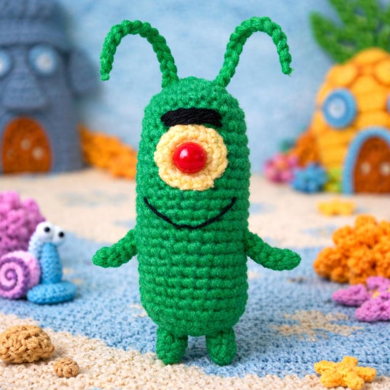 May include: A handmade green crochet Plankton character from SpongeBob SquarePants. The plush toy has a red eye, yellow and black details, and small arms and legs. Other crochet characters and a beach scene are in the background.