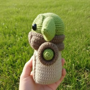 May include: A crocheted doll resembling a character with large ears and a distinctive face. The doll is mainly green, brown, and beige, with a round body and large, black eyes. The doll is held in a hand against a backdrop of green grass.