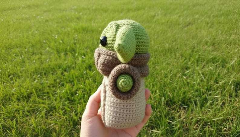 May include: A crocheted doll resembling a character with large ears and a distinctive face. The doll is mainly green, brown, and beige, with a round body and large, black eyes. The doll is held in a hand against a backdrop of green grass.