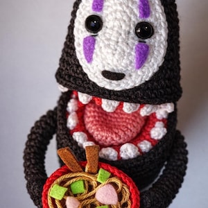 Puede incluir: Peluche de No-Face tejido a ganchillo de El Viaje de Chihiro, con una cara blanca, marcas moradas y ojos negros. Sostiene un cuenco rojo de ramen con fideos marrones, ingredientes verdes y rosas, y palillos marrones.