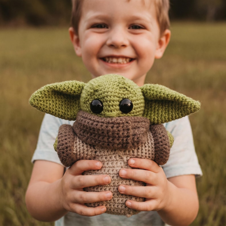 May include: A crocheted plush toy resembling a character with large ears and black eyes. The toy is mainly green, brown, and beige, and is held by a person. The background is blurred, suggesting an outdoor setting.