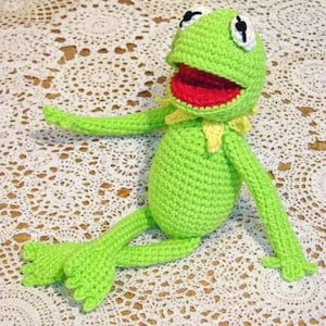 May include: A handmade green crochet frog doll with a wide open red mouth, white eyes, and yellow collar. The frog is sitting with its arms and legs extended. The doll is made of soft yarn and is a whimsical toy.